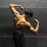Shirtless man flexing biceps against gray wall, showing muscular arms and torso